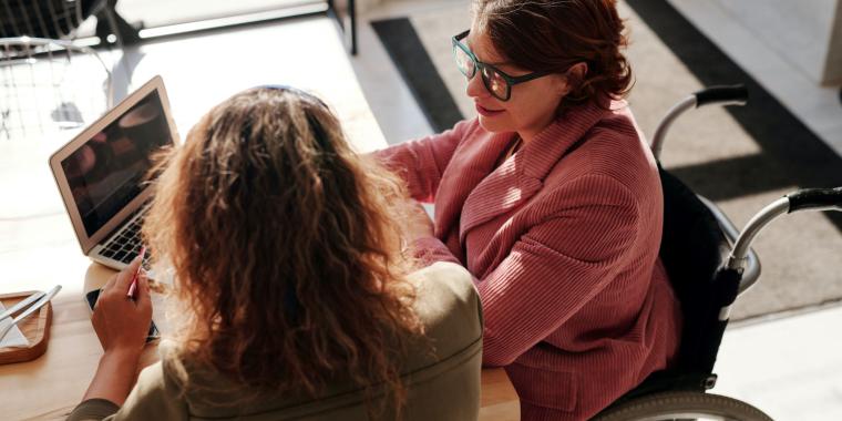Two women meeting. One is using a laptop, is in a wheelchair and is smiling. The other has her back to the camera.