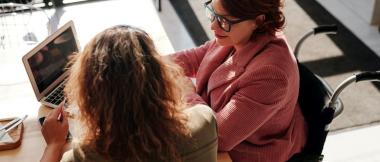 Two women meeting. One is using a laptop, is in a wheelchair and is smiling. The other has her back to the camera.